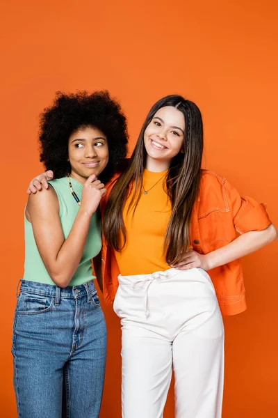 Smiling and stylish brunette teenager with colorful makeup hugging african american girlfriend in casual outfit isolated on orange, teen fashionistas with impeccable style concept, diverse races — Stock Photo
