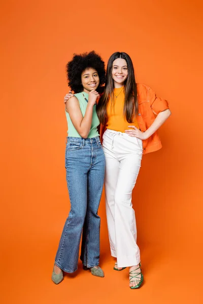 Full length of joyful brunette teen girl in stylish outfit hugging african american girlfriend with bright makeup on orange background, teen fashionistas with impeccable style concept — Stock Photo