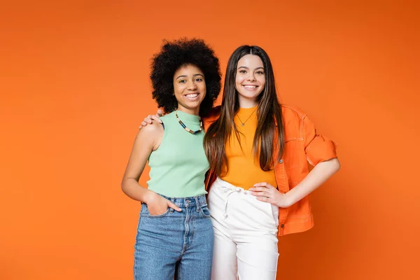 Positive and multiethnic girlfriends with bold makeup and trendy outfits hugging and looking at camera while posing isolated on orange, teen fashionistas with impeccable style concept — Stock Photo