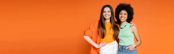 Cheerful interracial teen girlfriends with bold makeup and trendy clothes hugging and looking at camera isolated on orange, teen fashionistas with impeccable style concept, banner — Stock Photo