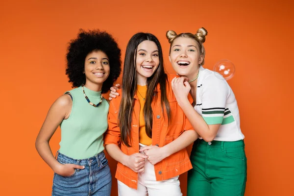 Positive and multiethnic teen girlfriends with bold makeup hugging and looking at camera near soap bubble on orange background, teen fashionistas with impeccable style concept — Stock Photo