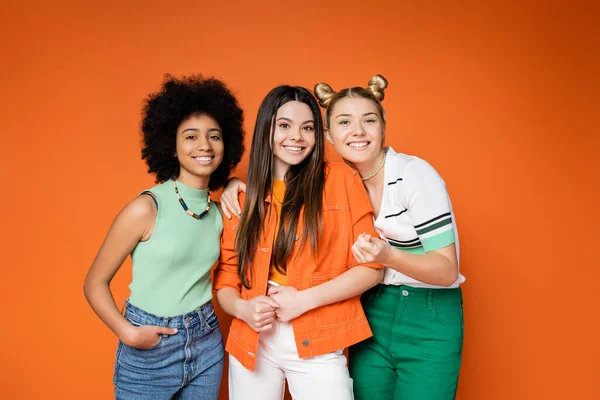 Novias adolescentes alegres y multiétnicas con maquillaje audaz que usan trajes casuales de moda que se abrazan y posan sobre un fondo naranja, adolescentes fashionistas con un concepto de estilo impecable - foto de stock