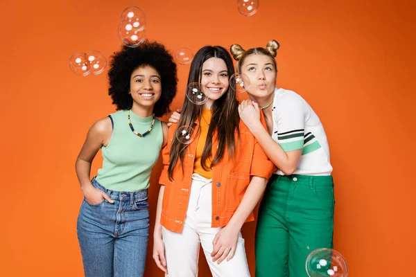 Positive and interracial girlfriends in stylish outfits hugging and posing near soap bubbles and standing on orange background, teen fashionistas with impeccable style concept, diverse races — Stock Photo