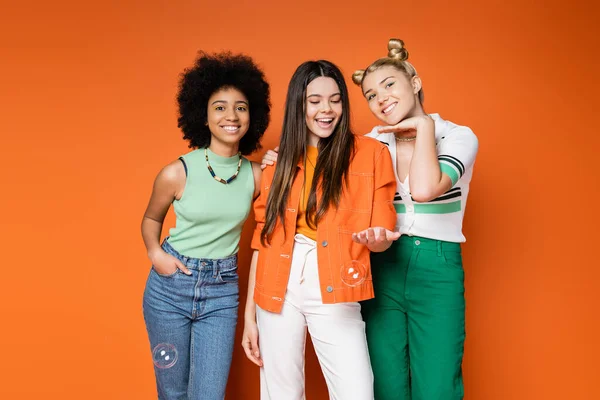 Fröhliche und trendige multiethnische Teenager-Mädchen mit fettem Make-up und lässiger Kleidung posieren mit Seifenblasen auf orangefarbenem Hintergrund, Teen-Fashionistas mit tadellosem Stil-Konzept — Stockfoto