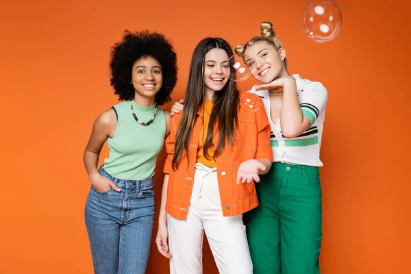Fröhliche und multiethnische Teen-Freundinnen in trendigen Casual-Outfits posieren und stehen neben Seifenblasen auf orangefarbenem Hintergrund, Teen-Fashionistas mit tadellosem Stylekonzept — Stockfoto