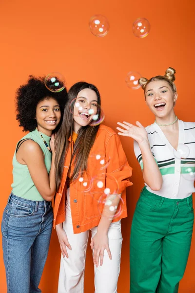 Seifenblasen in der Nähe positiver und interrassischer Teenager-Freundinnen in lässigen Outfits, die auf orangefarbenem Hintergrund in die Kamera schauen, Teen-Fashionistas mit tadellosem Stilkonzept — Stockfoto