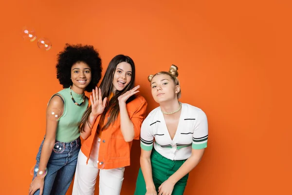 Novias multiétnicas y adolescentes con elegantes trajes casuales mirando a la cámara y posando cerca de burbujas de jabón mientras están de pie sobre fondo naranja, adolescentes fashionistas con un concepto de estilo impecable - foto de stock