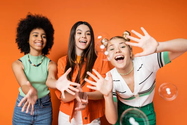 Захватывающие и веселые многонациональные девочки-подростки с смелым макияжем, смотрящие на мыльные пузыри, позируя и стоя на оранжевом фоне, подростки модницы с безупречной концепцией стиля — стоковое фото