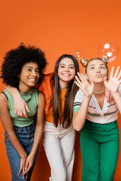 Fröhliche brünette Teenager umarmen stilvolle multiethnische Freundinnen und stehen in der Nähe von Seifenblasen auf orangefarbenem Hintergrund, multiethnische Teen-Fashionistas mit tadellosem Stilkonzept — Stockfoto