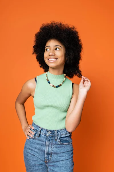 Stylish african american teenage girl in casual outfit and necklace touching hair, holding hand on hip and looking away isolated on orange, trendy teenage girl expressing individuality — Stock Photo
