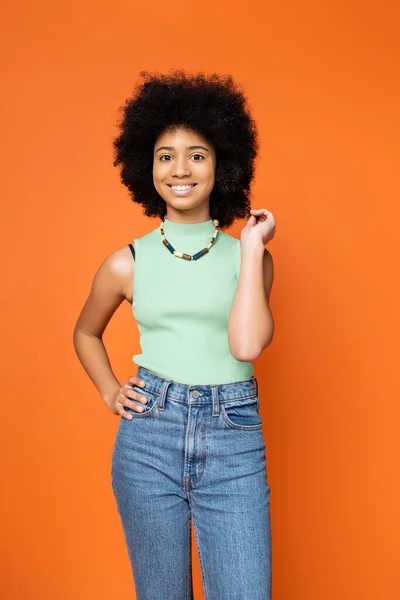 Chica adolescente afroamericana positiva con maquillaje audaz posando en un traje casual elegante y tocando el cabello mientras está de pie aislado en naranja, chica adolescente de moda que expresa individualidad - foto de stock