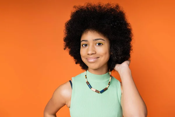 Retrato de adolescente afroamericano sonriente y elegante con maquillaje audaz usando collar y tocando el cabello mientras mira a la cámara aislada en naranja, chica adolescente de moda que expresa individualidad - foto de stock