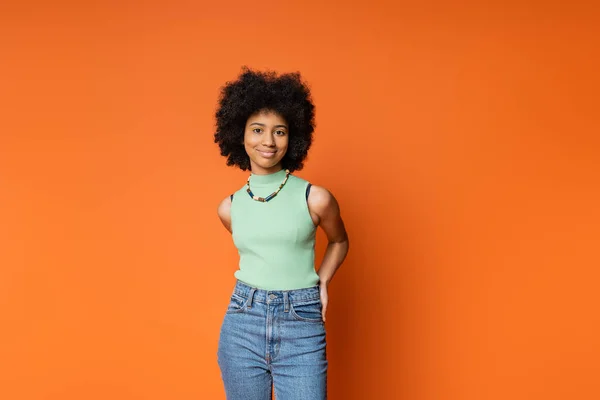 Smiling and stylish african american teenage girl in casual clothes with necklace and bold makeup posing and looking at camera isolated on orange, trendy teenage girl expressing individuality — Stock Photo
