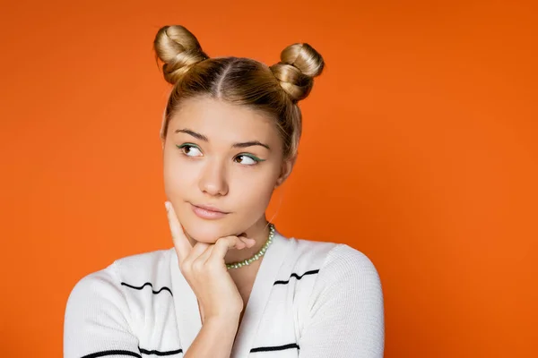 Portrait of pensive and stylish teenage girl with hairstyle and bold makeup looking away while posing in casual clothes isolated on orange, trendy teenage girl expressing individuality — Stock Photo
