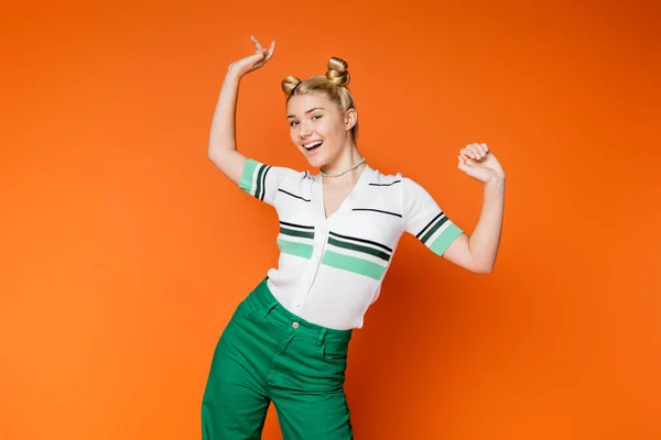 Aufgeregt und positiv blonde Teenager-Mädchen mit Frisur und buntem Make-up posiert in stilvollen lässigen Outfit und Blick in die Kamera auf orangefarbenem Hintergrund, modische und trendige Kleidung — Stockfoto