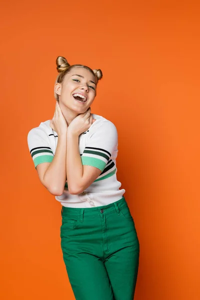 Porträt eines fröhlichen blonden Teenie-Mädchens mit stilvoller Frisur und kühnem Make-up, das den Hals berührt und in die Kamera schaut, während es isoliert auf orangefarbener, modischer und trendiger Kleidung posiert — Stockfoto