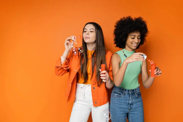 Stylish brunette teenage girl blowing soap bubbles near cheerful african american girlfriend with bold makeup while standing together on orange background, fashionable and trendy clothes — Stock Photo