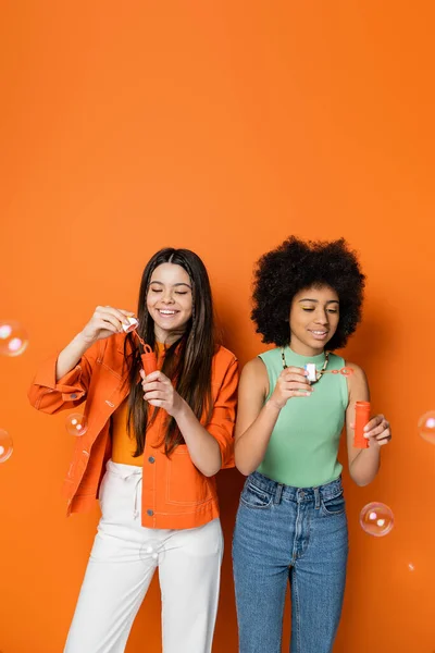 Smiling brunette and african american teenage girlfriends with bold makeup in casual outfits holding soap bubbles and standing on orange background, fashionable and trendy clothes — Stock Photo