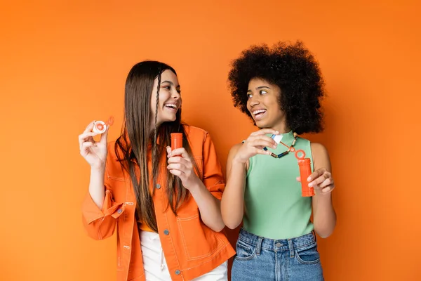 Positive und trendige multiethnische Teenager mit kühnem Make-up und lässigen Outfits, die einander anschauen, während sie Seifenblasen halten und auf orangefarbenem Hintergrund stehen, modische und trendige Kleidung — Stockfoto