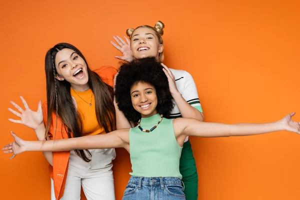 Chicas adolescentes alegres y multiétnicas con maquillaje colorido posando juntos en trajes casuales y mirando a la cámara mientras están de pie sobre fondo naranja, ropa de moda y de moda - foto de stock