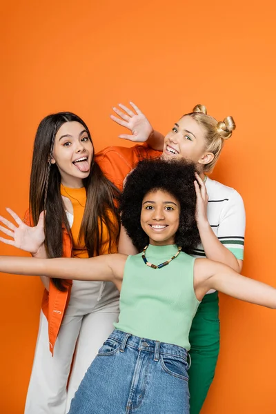 Jovens namoradas alegres e multiétnicas com maquiagem colorida posando em roupas casuais e olhando para a câmera juntas enquanto estão isoladas em roupas laranja, na moda e na moda — Fotografia de Stock