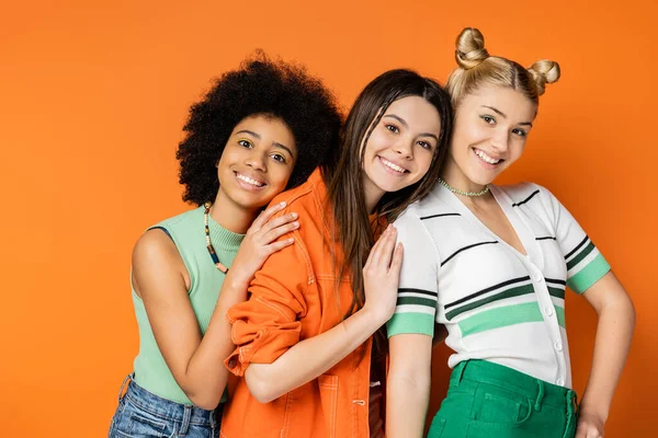 Живые многоэтнические подростки с смелым макияжем в стильной повседневной одежде и обнимая блондинку, стоя вместе на оранжевом фоне, модной и модной одежде — стоковое фото