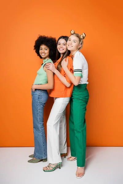 Comprimento total de adolescentes multiétnicos na moda com maquiagem ousada vestindo roupas casuais e abraços enquanto estão ao lado um do outro em fundo laranja, poses elegantes e confiantes — Fotografia de Stock