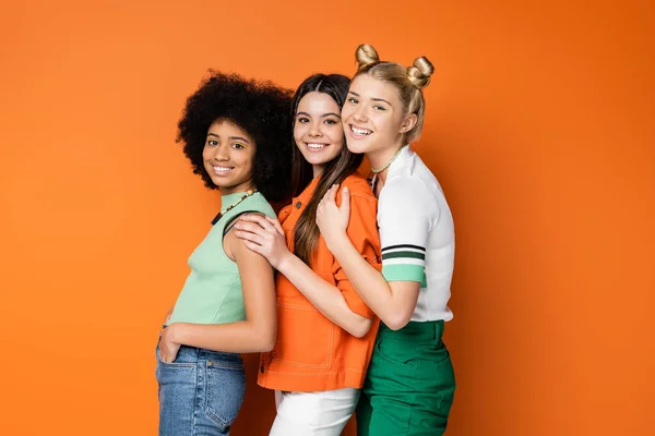 Modernas y sonrientes novias adolescentes multiétnicas con maquillaje audaz que usan atuendos casuales mientras posan y miran a la cámara en un fondo naranja, poses elegantes y confiadas - foto de stock