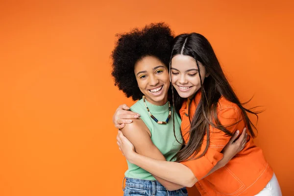 Menina americana africana alegre e adolescente com maquiagem colorida abraçando namorada morena em roupa casual e olhando para a câmera no fundo laranja, poses elegantes e confiantes — Fotografia de Stock