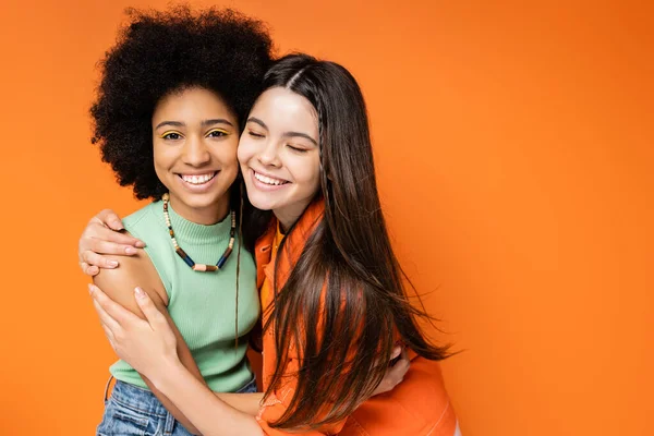 Joyeux adolescent brune en tenue décontractée étreignant petite amie afro-américaine à la mode avec un maquillage coloré et debout ensemble sur fond orange, poses élégantes et confiantes — Photo de stock