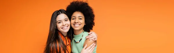 Lächelnder brünetter Teenager in lässiger Kleidung umarmt afrikanisch-amerikanische Freundin mit kühnem Make-up und blickt isoliert in die Kamera auf orangefarbenen, stilvollen und selbstbewussten Posen, Banner — Stockfoto