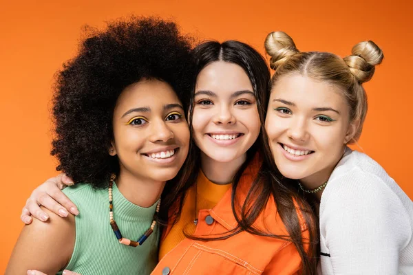 Porträt eines lächelnden Teenagers mit buntem Make-up, der multiethnische Freundinnen umarmt und gemeinsam in die Kamera schaut, während er isoliert auf orangefarbenen, stilvollen und selbstbewussten Posen posiert — Stockfoto