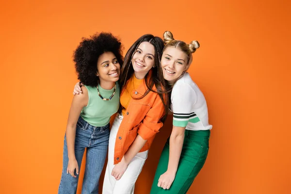 Позитивные и стильные многоэтнические девушки-подростки с красочным макияжем, обнимающие брюнетку и позирующие вместе на оранжевом фоне, модные и стильные прически — стоковое фото