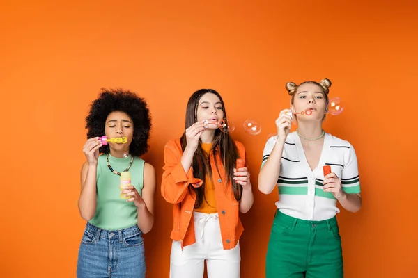 Multiethnic teenage girlfriends with bold makeup and casual outfits blowing soap bubbles and spending time together while standing on orange background, trendy and stylish hairstyles — Stock Photo