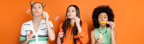 Interracial teenage girlfriends with colorful makeup wearing casual clothes while blowing soap bubbles and spending time on orange background, trendy and stylish hairstyles, banner — Stock Photo