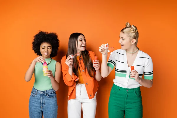 Позитивні і модні дівчата-підлітки з барвистим макіяжем в повсякденному вбранні і тримають мильні бульбашки біля афроамериканської дівчини на помаранчевому фоні, модні і стильні зачіски — стокове фото