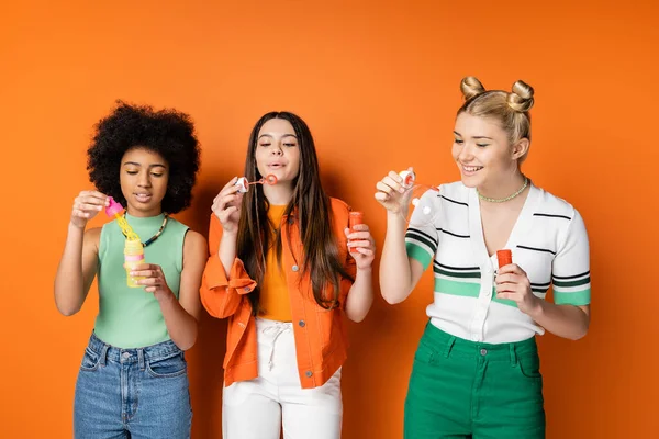 Cheerful blonde teenage girl in casual clothes holding soap bubbles near multiethnic girlfriends with colorful makeup while standing on orange background, trendy and stylish hairstyles — Stock Photo