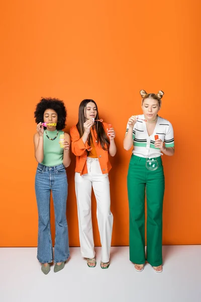 Full length of trendy and multiethnic teenage girlfriends with colorful makeup wearing casual outfits while blowing soap bubbles and standing on orange background, trendy and stylish hairstyles — Stock Photo