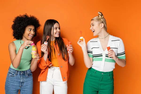 Positive blonde teenager with bold makeup holding soap bubbles and talking to multiethnic girlfriends in casual clothes while standing on orange background, trendy and stylish hairstyles — Stock Photo