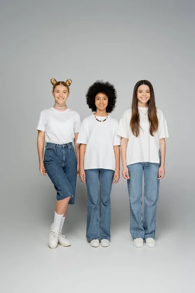 Longitud completa de adolescentes multiétnicos animados y elegantes en camisetas blancas y pantalones vaqueros mirando juntos a la cámara mientras están de pie uno al lado del otro sobre un fondo gris, looks modernos y elegantes - foto de stock