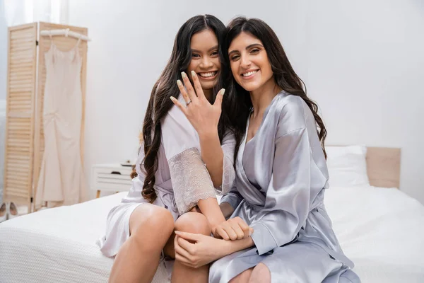 Happy asian woman and her friend sitting on bed, showing engagement ring, happiness, slumber party, silk robes, best friends, bride with her bridesmaid, brunette hair, cultural diversity, multiracial — Stock Photo