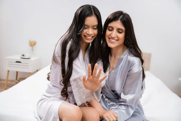 Happy asian woman and her friend sitting on bed, showing engagement ring, happiness, bridal party, silk robes, best friends, bride with her bridesmaid, brunette hair, cultural diversity, multiracial — Stock Photo