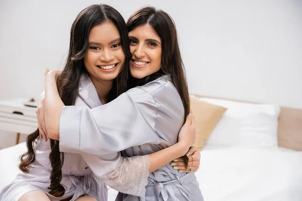 Happy asian woman and her friend sitting on bed, hugging each other, bridal party, silk robes, best friends, bride with her bridesmaid, brunette hair, cultural diversity, multiracial, pre-wedding — Stock Photo