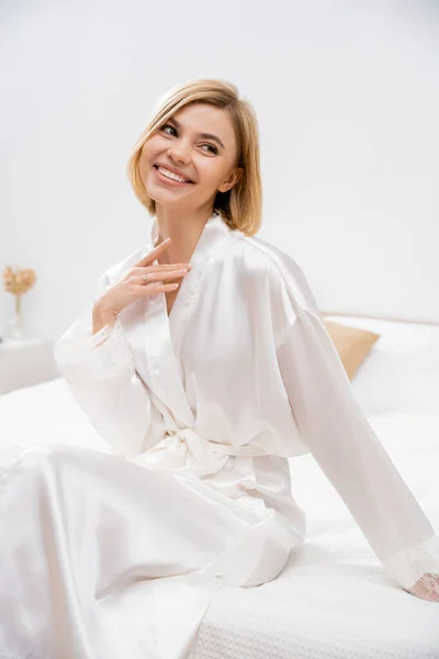 Cheerful bride with blonde hair sitting in white silk robe on bed, looking away, smiling, wedding planning, young woman, beautiful, excitement, feminine, blissful — Stock Photo