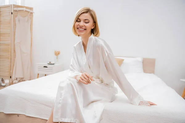 Cheerful bride with blonde hair sitting in white silk robe on bed, smiling, wedding planning, young woman, beautiful, excitement, feminine, blissful, preparation, pre-wedding — Stock Photo