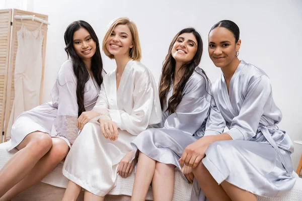 Bridal party, silk robes, best friends, bride with her multicultural bridesmaids, brunette and blonde women, cultural diversity, happy girlfriends sitting on bed, positivity, looking at camera — Stock Photo
