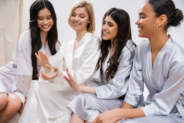 Bridal party, silk robes, best friends, bride with her multicultural bridesmaids looking at engagement ring, brunette and blonde, cultural diversity, sitting on bed, positivity, wedding preparations — Stock Photo