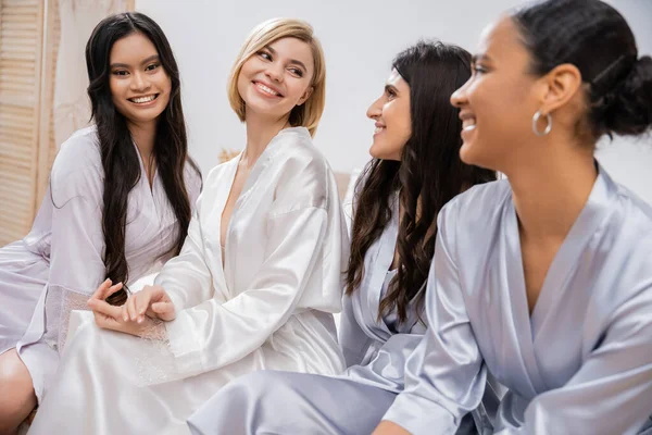 Bridal party, silk robes, female friends, bride with her multicultural bridesmaids, brunette and blonde women, cultural diversity, happy girlfriends sitting on bed, positivity, wedding preparations — Stock Photo