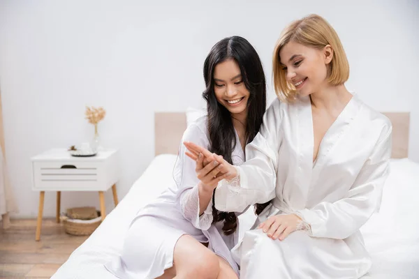 Happy blonde bride and her asian bridesmaid sitting on bed, showing engagement ring, happiness, bridal party, silk robes, brunette and blonde women, diversity, multicultural female friends — Stock Photo