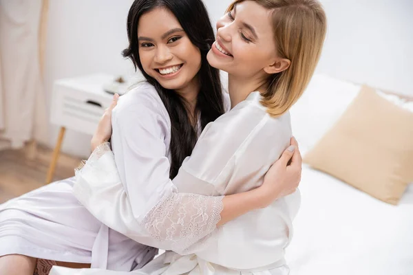 Happy blonde bride hugging with her asian bridesmaid, happiness, bridal party, silk robes, brunette and blonde women, diversity, multicultural best friends, excitement, friendship goals — Stock Photo